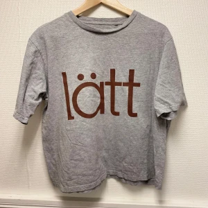 Grå t-shirt med tryck - Säljer en grå t-shirt med brunt tryck 'lätt' på framsidan. T-shirten har en boxig passform och är kortärmad. Perfekt för en avslappnad stil.