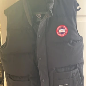 Svart dunväst från Canada Goose - Säljer en svart dunväst från Canada Goose med klassisk logga på bröstet. Västen har praktiska fickor och knäppning framtill. Perfekt för kyliga dagar.