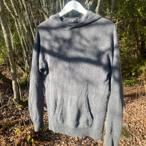 Grå hoodie i silke och kashmir - Säljer en lyxig grå hoodie från Silk & Cashmere, tillverkad i en mjuk blandning av silke och kashmir. Tröjan har en klassisk design med huva och ribbade muddar. Perfekt för en stilren och bekväm look. Köpte i tyskland för 2400kr