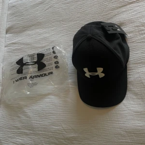 Under Armour keps - Säljer en snygg svart keps från Under Armour i nytt skick. Kepsen är i storlek M/L men har en perfekt passform på de flesta då den är i stretchigt material. Perfekt för både vardag och träning!