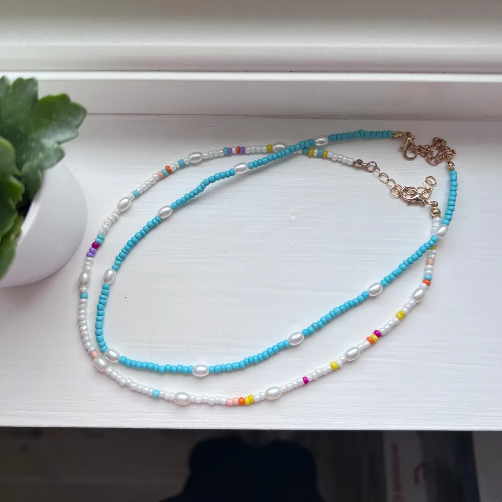 Snygga halsband med små pärlor i turkos, vitt och flera färger. Halsbanden har justerbara kedjor i guld och är perfekt för att ge en färgklick till din outfit. Pärlorna är arrangerade i ett lekfullt mönster med inslag av rosa, gul och orange.. Asusteet.