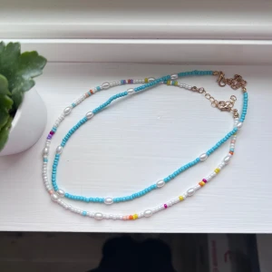 Färgglada pärlhalsband - Snygga halsband med små pärlor i turkos, vitt och flera färger. Halsbanden har justerbara kedjor i guld och är perfekt för att ge en färgklick till din outfit. Pärlorna är arrangerade i ett lekfullt mönster med inslag av rosa, gul och orange.