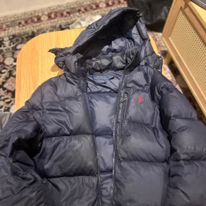 Mörkblå dunjacka från Ralph Lauren - Säljer en snygg mörkblå dunjacka från Ralph Lauren med huva och dragkedja. Jackan har en röd logotyp på bröstet och en praktisk bröstficka med dragkedja. Perfekt för kyliga dagar!( tröjan är en xs men sitter som s/M har M i kläder och den passar med marginal) ny 6000kr kvitt finns men borta (från NK)