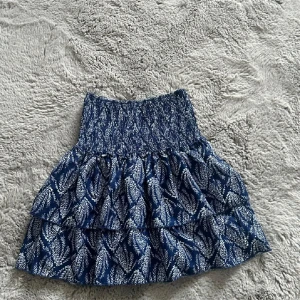 Blå mönstrad kjol - Snygg blå kjol med vitt mönster och volanger. Den har en elastisk midja för bekväm passform. Perfekt för en somrig look!