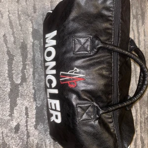Svart väska från Moncler - Snygg svart väska från Moncler i skinn med vit logga och detaljer. Väskan har en dragkedja och bärhandtag, perfekt för att bära med sig det nödvändiga. Den har en stilren design med vita kanter och en praktisk storlek.