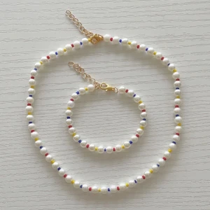 Pärlset halsband & armband färgglada detaljer - Säljer ett pärlset med halsband och armband. Båda har vita pärlor med små inslag av röda, blå och gula pärlor. Kedjor och lås är i guldton. Perfekt för dig som gillar färgglada accessoarer med en lekfull touch.