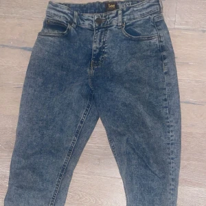 Blå Lee jeans high waist 12-13Y - Snygga blå Lee jeans med high waist och klassisk femficksdesign. Jeansen har en tvättad look och raka ben, perfekt för dig som gillar en avslappnad men trendig stil. Tydlig Lee-logga bak på linningen och slitstarka sömmar.