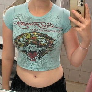 Ed Hardy BABY-T - Säljer en turkos baby-t från Ed Hardy med ikoniskt tigertryck framtill och glittriga detaljer. Toppen har korta ärmar, tight passform och tryckt logga både fram och bak. Perfekt för dig som gillar statement-plagg och streetstyle.
