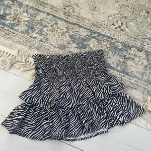 Zebramönstrad volangkjol svart/vit - Supersöt kort kjol med zebramönster i svart och vitt. Kjolen har smockad midja och dubbla volanger som ger en riktigt flowy och lekfull look. Perfekt för dig som vill sticka ut med en trendig och djurinspirerad stil.