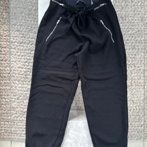 Svarta byxor med dragkedjor - Svarta byxor med elastisk midja och snörning, samt dragkedjor vid fickorna framtill. Byxorna har resår vid bensluten för en skön och avslappnad passform.