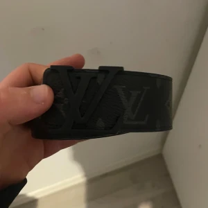 Svart  skärp i skinn - Snyggt svart  bälte i skinn med klassiskt monogram i mörkgrått och svart. Bältet har en bred design och en mattsvart späne framtill. Insidan är ljusbrun och bältet har exklusiv känsla, perfekt för att lyfta din outfit. Man får me så man kan göra större hål 