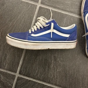 Blåa Vans Old Skool sneakers - Säljer ett par klassiska Vans Old Skool sneakers i blått med vita detaljer och snörning. Skorna har vit sula och den ikoniska sidoremsan. Ovandelen är i mocka och textil, perfekt för dig som gillar streetstyle.