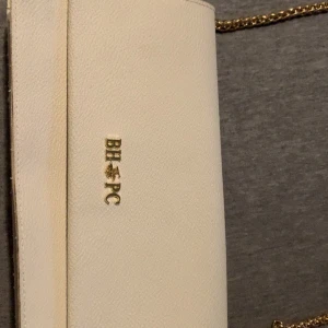 Beige axelväska från BHPC med kedja - Snygg beige axelväska från BHPC med guldfärgad kedjerem och logga framtill. Väskan är i skinnimitation och har flera fack, inklusive ett med dragkedja. Perfekt storlek för det viktigaste och stilren design som lyfter din outfit.