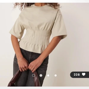  korsett-topp med kort ärm - Trendig beige topp med korsettliknande midja och korta ärmar. Toppen har en markerad, figurnära passform över midjan med snygga sömdetaljer som ger en cool siluett. Perfekt att styla med jeans eller kjol för en modern look.