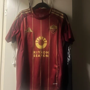 AS Roma röd matchtröja Adidas - Snygg AS Roma fotbollströja från Adidas i rött med guldiga detaljer och ränder. Tröjan har klubbmärke på bröstet, guldiga Adidas-loggor och 'Riyadh Season' tryckt framtill. Tillverkad i lätt funktionsmaterial med korta ärmar och klassisk passform.