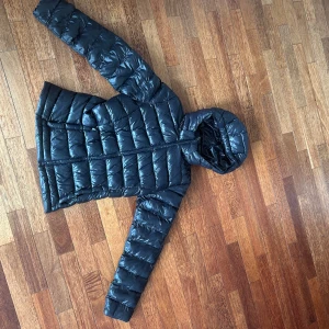  Jacka  - Snygg svart pufferjacka med huva och dragkedja framtill. 