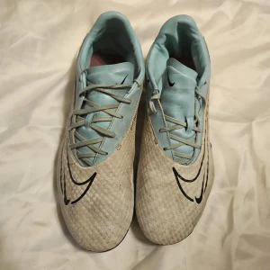 Nike Phantom GX fotbollsskor ljusblå - Nike Phantom GX fotbollsskor i en snygg ljusblå och beige färgkombination. Skorna har snörning, svart Nike-logga och ovandel i syntetmaterial med textur för bättre bollkontroll. Yttersulan har dobbar för gräsplan. Perfekta för dig som vill sticka ut på planen.
