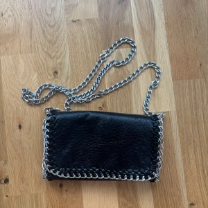 Svart tiamo väska - Cool svart axelväska från TIAMO i skinnimitation med  silverfärgade kedjor runt kanterna och som axelrem. Väskan har magnetlås, innerfack med dragkedja och snygga flätade detaljer längs sidorna. Lite sliten inuti men inget som syns när man har den stängd! Skriv för mer frågor eller bilder, tar emot prisförslag!🩷
