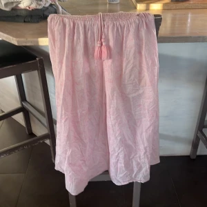 Ljusrosa långkjol från Gina Tricot - Supersöt ljusrosa långkjol från Gina Tricot med diskret mönster och smock i midjan. Kjolen har en dragsko med tofsar framtill och är gjord i ett tunt, luftigt bomullstyg. Perfekt för en chill sommardag eller när du vill känna dig extra söt.