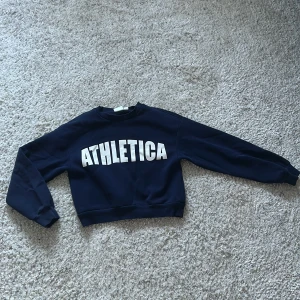 Mörkblå sweatshirt Gina Tricot - Mörkblå sweatshirt från Gina Tricot med stort vitt ATHLETICA-tryck. Tröjan har rund hals, långa ärmar och ribbade muddar. Materialet är mjukt och skönt. Använt 2-3 gg, inga defekter! 