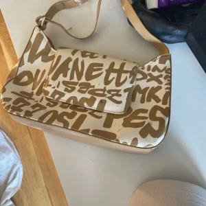 Beige axelväska med grafiskt tryck - Cool beige axelväska med brunt grafiskt textmönster och justerbar axelrem. Väskan har flera fack dock så är magnetlåset framtill trasigt men det går fortfarande att stänga. Materialet är syntet och insidan är fodrad i beige tyg. Perfekt för dig som gillar unika accessoarer.