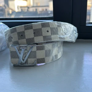 L bälte - Snyggt skärp  med klassiskt rutigt mönster i beige och grått. Bältet har ett stort, silverfärgat spänne framtill och är gjort i skinn. Perfekt accessoar för att lyfta din outfit med en exklusiv touch.
