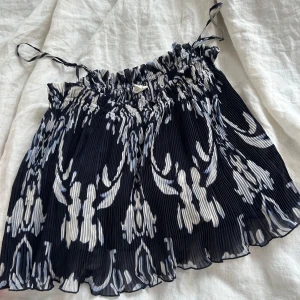 Svartvit plisserad linnetopp - Supersöt plisserad linnetopp med svart och vitt abstrakt mönster. Toppen har smala knytband vid axlarna och en luftig, flowy känsla. Perfekt för varma dagar när du vill ha något lätt och snyggt.