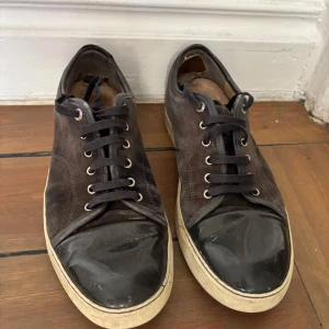 Lanvin skor - Snygga svarta sneakers från Lanvin med ovandel i mocka och glansig tå i svart lack. Klassisk rund tå, svarta snören och vit sula ger en stilren look. Perfekta för dig som gillar exklusiva och tidlösa sneakers med en twist. Liten defekt under båda skorna men vatten läcker inte in. 