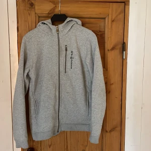 Grå hoodiejacka från Sail Racing - Ljusgrå hoodiejacka från Sail Racing med hel dragkedja, huva och broderad logga. Jackan har en bröstficka med dragkedja och två sidofickor. Her en liten fett fläck bredvid bröstfickan, inte hjätte synlig.