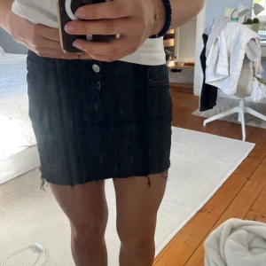 Svart jeanskjol - Säljer en svart kort jeanskjol med rå, fransig kant nertill. Kjolen har klassisk femficksdesign och stängs med knapp och dragkedja framtill. Perfekt för dig som gillar en enkel men edgy stil.