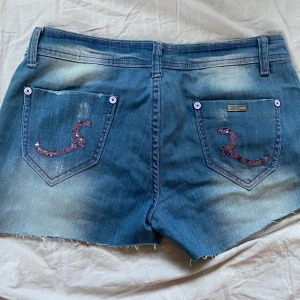 Blå jeansshorts med paljettdetaljer - Lågmidjade blå jeansshorts från Miss Sixty med råa kanter och lila paljettdetaljer på fickorna. Shortsen har lila sömmar, slitningar och coola knappar med lila text. 