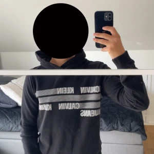 Svart Calvin Klein Jeans hoodie - Svart hoodie från Calvin Klein Jeans med vit logga och ränder över bröstet. Klassisk huva, långa ärmar och ribbade muddar. Perfekt för en chill och stilren look. Mjuk bomullskänsla och snygg passform.