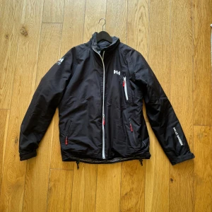 Svart vindjacka från Helly Hansen - Svart vindjacka från Helly Hansen med vit dragkedja och röda detaljer på fickorna. Jackan har hög krage, tre fickor med dragkedja och justerbar nederkant. Perfekt för blåsiga dagar och snygg logga på bröstet och ärmen.