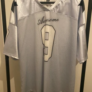 Silver fotbollströja Shein nr 9 - Snygg silverfärgad fotbollströja från Shein i storlek L. Tröjan har svarta detaljer, nummer 9 på ryggen och texten 'Awesome'. Tillverkad i glansigt, syntetiskt material med meshpanel framtill och svarta stjärnor på axlarna. Perfekt för dig som gillar sportig stil.