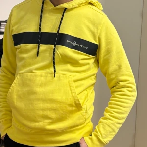 Gul hoodie från Sail Racing - Gul hoodie från Sail Racing med svart horisontell rand och logga över bröstet. Tröjan har huva med svarta snören och en stor magficka. Perfekt för dig som gillar sportig och färgstark stil.