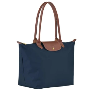Blå Longchamp axelväska i nylon - Stilren marinblå axelväska från Longchamp med bruna skinnhandtag och lock. Väskan är rymlig med dragkedja upptill och guldfärgade detaljer. Tillverkad i slitstarkt nylon med klassisk design och insida i svart tyg. Perfekt för dig som vill ha både stil och funktion. Väskan är precis som nyskick!
