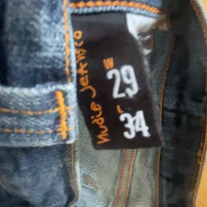 Blå  jeans från Nudie  - Säljer ett par klassiska blå skinny jeans från Nudie Jeans med snygga orangea sömmar och broderade bakfickor. Jeansen har normal passform i midjan och är tillverkade i mjukt denimtyg.
