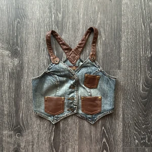 Jeansväst med bruna skinn-detaljer - Unik croppad jeansväst från Miss Sixty med bruna skinnremmar och fickor. Västen har knappar framtill, coola sömmar och detaljer i brunt skinn både fram och bak. Perfekt för dig som vill sticka ut med en edgy look. Liten i storlek mer som xs 