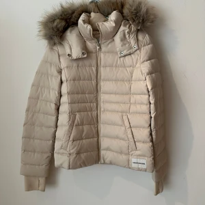 Beige dunjacka från Calvin Klein Jeans Strl.S - Snygg beige dunjacka från Calvin Klein Jeans med avtagbar huva och fuskpäls. Jackan har quiltad design, dragkedja framtill och två fickor med fleece på insidan. Perfekt för kalla dagar med sin varma dun- och fjäderfyllning. Stilsäker och enkel att matcha till det mesta. 