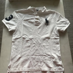 Vit pikétröja Polo Ralph Lauren - Vit pikétröja från Polo Ralph Lauren med klassisk krage, korta ärmar och broderad marinblå logga på bröstet. Slim fit-modell med knappar framtill och stor siffra 3 på ena ärmen. Tillverkad i bomull för en skön känsla. Pikétröjan hade ett hål på axeln men är lagad. 