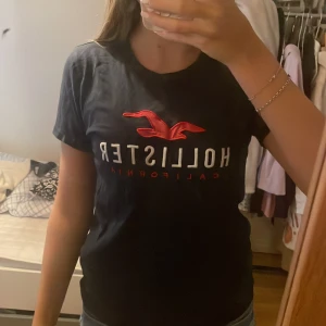 Mörkblå Hollister t-shirt med röd logga - Super snygg original mörkblå t-shirt från Hollister med klassisk röd fågellogga och vit text på bröstet. Modellen är kortärmad med rund halsringning och normal passform. Perfekt för en chill och stilren look.