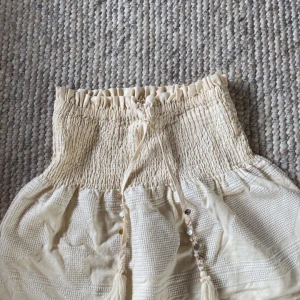 Beige kortkjol med snäckdetaljer från Zara - Supersöt beige kortkjol från Zara med smockad midja och knytband dekorerade med snäckor och pärlliknande detaljer. Kjolen har en strukturerad yta och volangkant upptill, perfekt för en chill sommarlook.