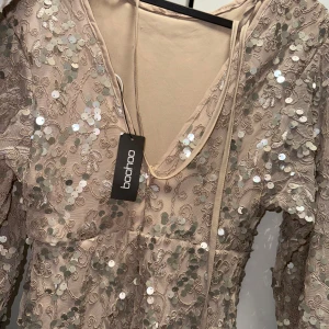 Champagnefärgad paljettklänning Boohoo - Oanvänd festklänning.  Champagnefärgad långklänning från Boohoo med v-ringning och långa ärmar. Klänningen är täckt av glittriga paljetter och har broderade detaljer över hela tyget. Perfekt för dig som vill sticka ut med en elegant och glittrig look.