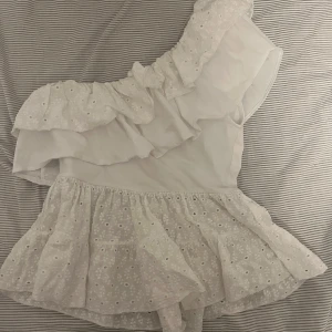 Vit omlott topp med volang från Zara - Supersöt vit omlott topp från Zara med volanger och broderade blommor. Toppen är oneshoulder och har en luftig, ärmlös design med broderad spets och volangdetaljer. Perfekt för sommarens alla tillfällen!