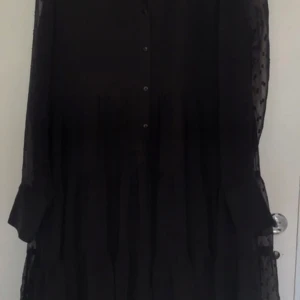 Svart långklänning från Gina Tricot - Säljer en svart medel långklänning från Gina Tricot med transparenta ärmar och diskret rutigt mönster. Klänningen har knappar framtill och är tillverkad i ett lätt, flowy material som ger en snygg siluett. Perfekt för dig som gillar stilrena och trendiga plagg.