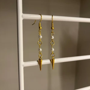 Guldiga örhängen med pärlor och spikes - Långa örhängen i guldton med kedjor, små vita pärlor och spetsiga spikes längst ner. Snygg kontrast mellan de eleganta pärlorna och de coola konformade detaljerna. Perfekta för att ge din look lite edge. Handgjort 