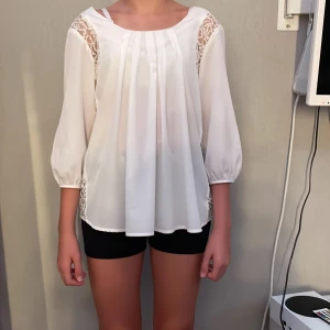 Vit blus med spetsdetaljer - Superfin vit blus i lätt och flowy material med veck framtill och spetsdetaljer längs axlar och sidor. Blusen har trekvartsärmar med resår vid ärmslut och rund halsringning. Perfekt för dig som gillar en romantisk och stilren look.