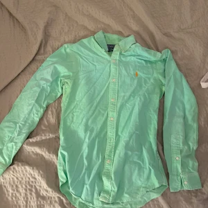 Mintgrön skjorta från Polo Ralph Lauren - Fräsch mintgrön skjorta från Polo Ralph Lauren i slim fit. Klassisk design med knappar hela vägen, lång ärm och ikonisk broderad logga i orange på bröstet. Skjortan är tillverkad i mjuk bomull och har en snygg krage.
