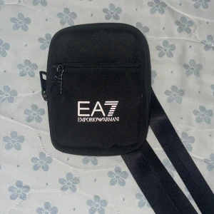 Svart axelväska EA7 Emporio Armani - Säljer en svart axelväska från EA7 Emporio Armani med vit logga framtill. Väskan har justerbar rem, dragkedja och ett smidigt fack på framsidan. Perfekt storlek för mobil och småprylar. Stilren och enkel design som passar till det mesta.