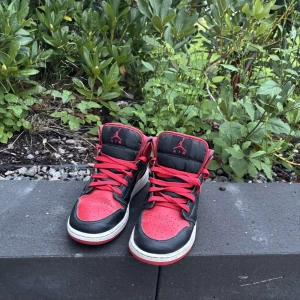 Nike Air Jordan 1 Bred Highs - Säljer ett par ikoniska Nike Air Jordan 1 i färgen Bred med svart och rött skinn, röda snören och klassisk vit sula. Skorna har högt skaft och den klassiska swoosh-loggan på sidan. Perfekta för dig som vill sticka ut med en tidlös sneaker.